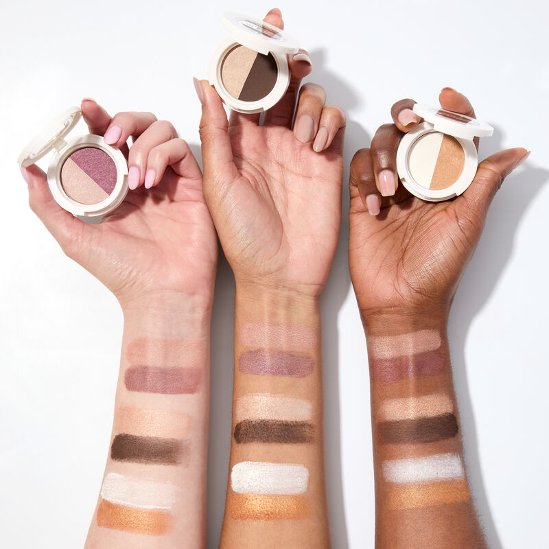 Swatches of special edition Mary Kay At Play Eye Shadow Duo in Rosy Plum, Choco Espresso and Gold Star on light to deep-skinned arms holding compacts