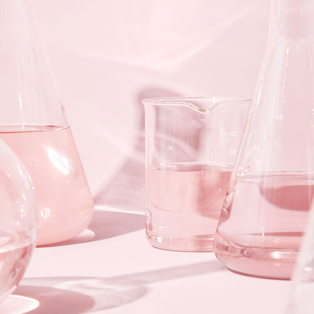 Vasos de vidrio transparentes con l&iacute;quido incoloro sobre una superficie rosa, reflejando una suave iluminaci&oacute;n rosada.