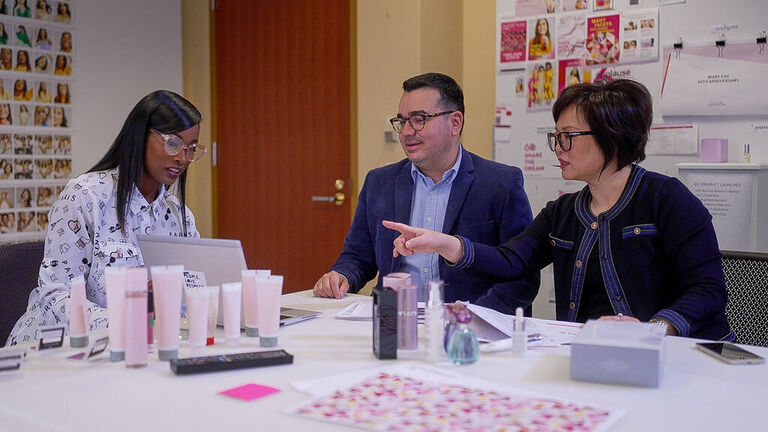 Tres colaboradores de Mary Kay trabajando en equipo alrededor de una mesa, con productos para el cuidado de la piel, una laptop y bocetos de dise&ntilde;o.