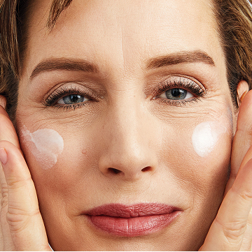 Mujer sonriendo a la c&aacute;mara mientras aplica la Loci&oacute;n Facial Regeneradora C + E TimeWise&reg; en ambas mejillas.