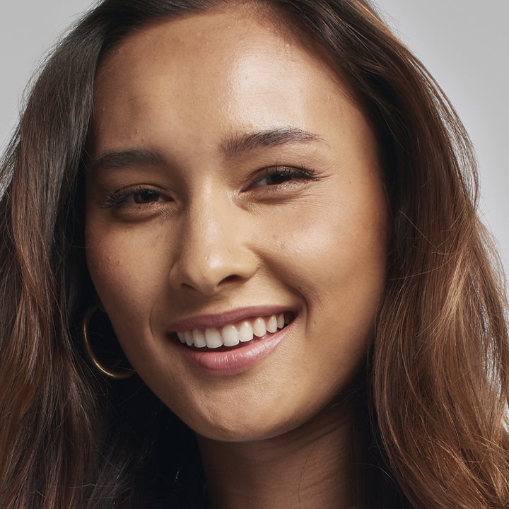 Modelo de cabello casta&ntilde;o sonriendo a la c&aacute;mara mientras usa su tono de base de acabado luminoso.